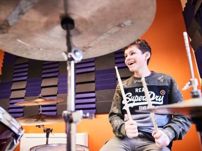 A boy wearing a Superdry jumper is playing the drums. He has a big smile on his face